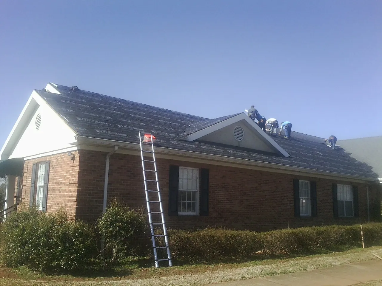 Crew tearing shingles off a brick ranch roof with a ladder set — Metal Roof Repair work by our crew in Clyman