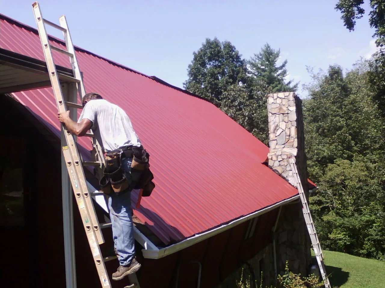 Roofer on a ladder beside a red metal roof and stone chimney — Soffit & Fascia Repair by Roofing Brothers in Clyman